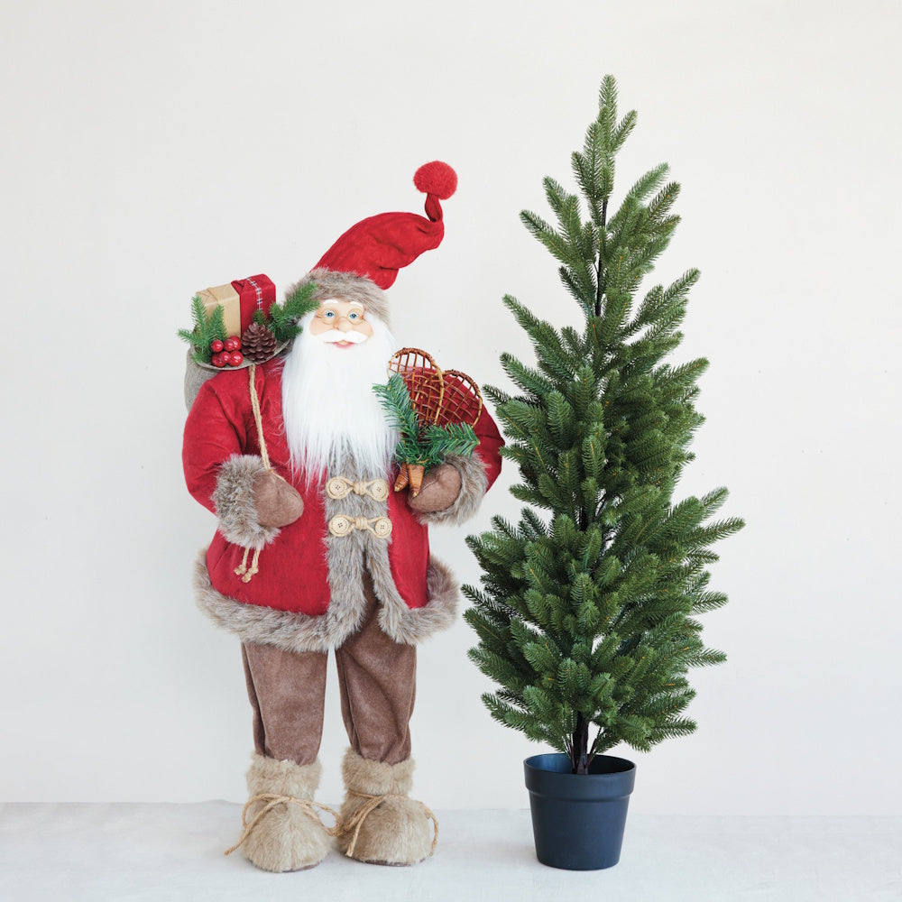 A fabric Santa figurine holding snowshoes next to a potted evergreen tree.