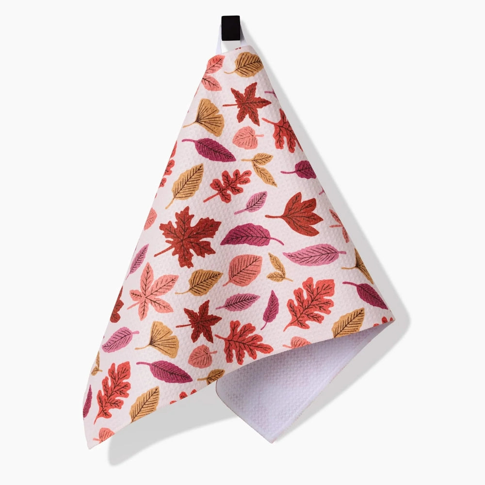 A hanging white kitchen towel with a waffle texture features a pattern of colorful, scattered autumn leaves.