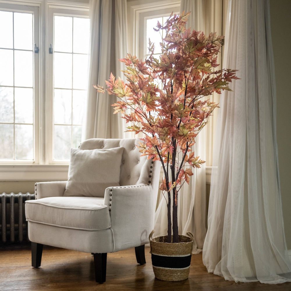 Faux autumn maple tree in a woven basket next to a cream armchair in front of a bright window.