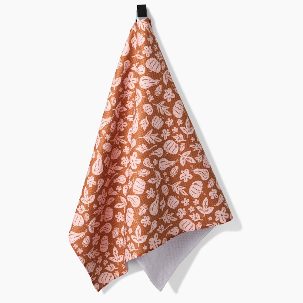A brown kitchen tea towel hanging up, featuring a light pink pattern of pumpkins, gourds, leaves, and flowers.
