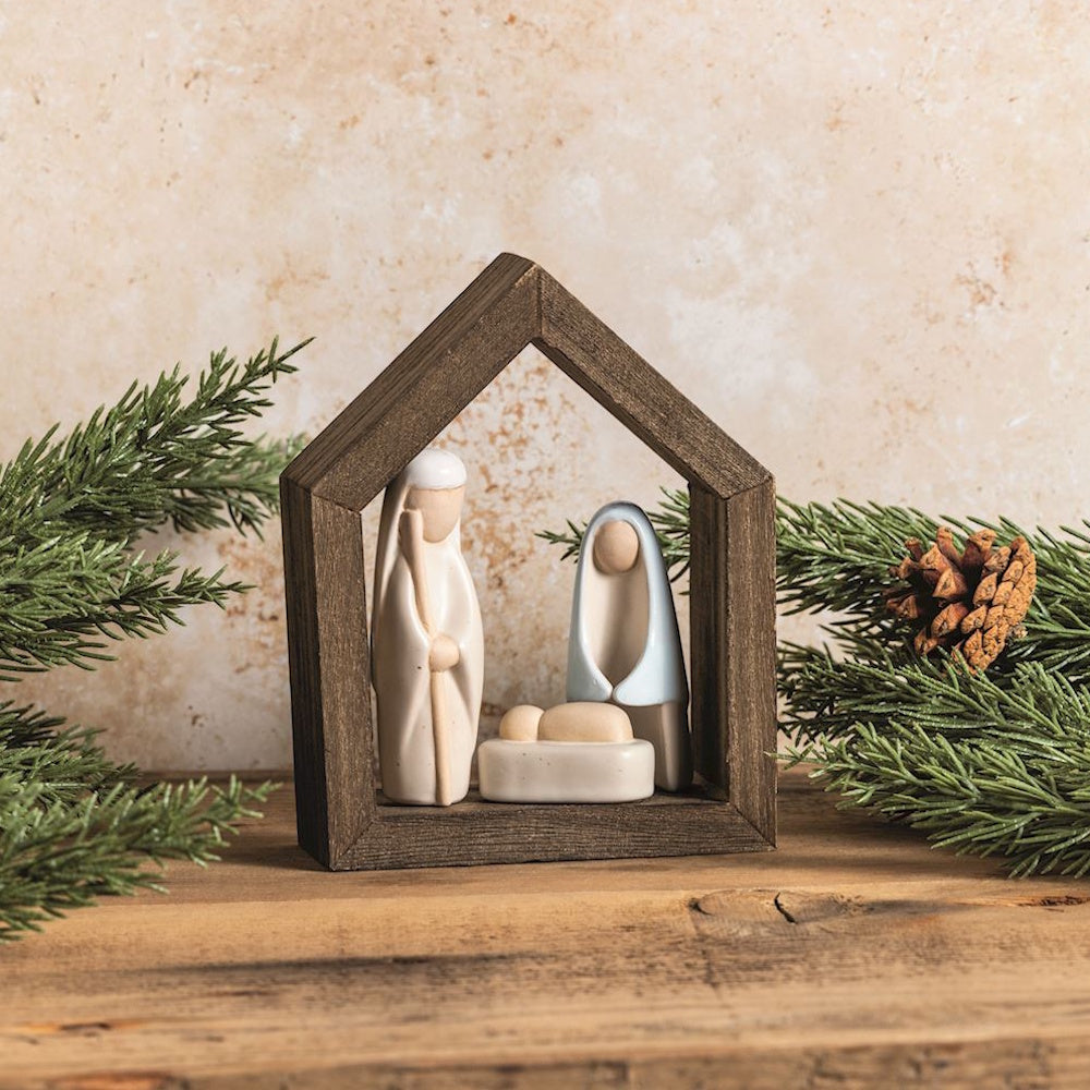 Minimalist ceramic Nativity scene set inside a dark wooden house frame surrounded by pine greenery.