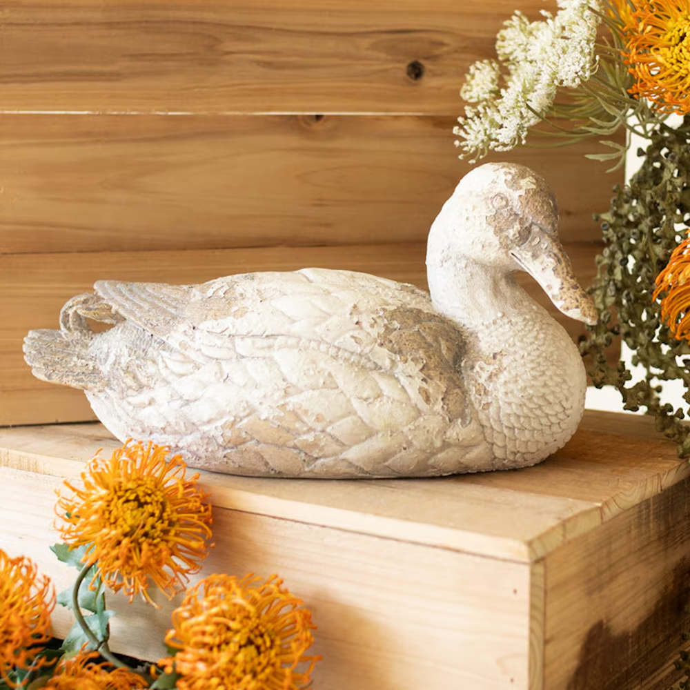 Rustic pintail duck statue resting on a wooden crate next to orange and white floral arrangements.
