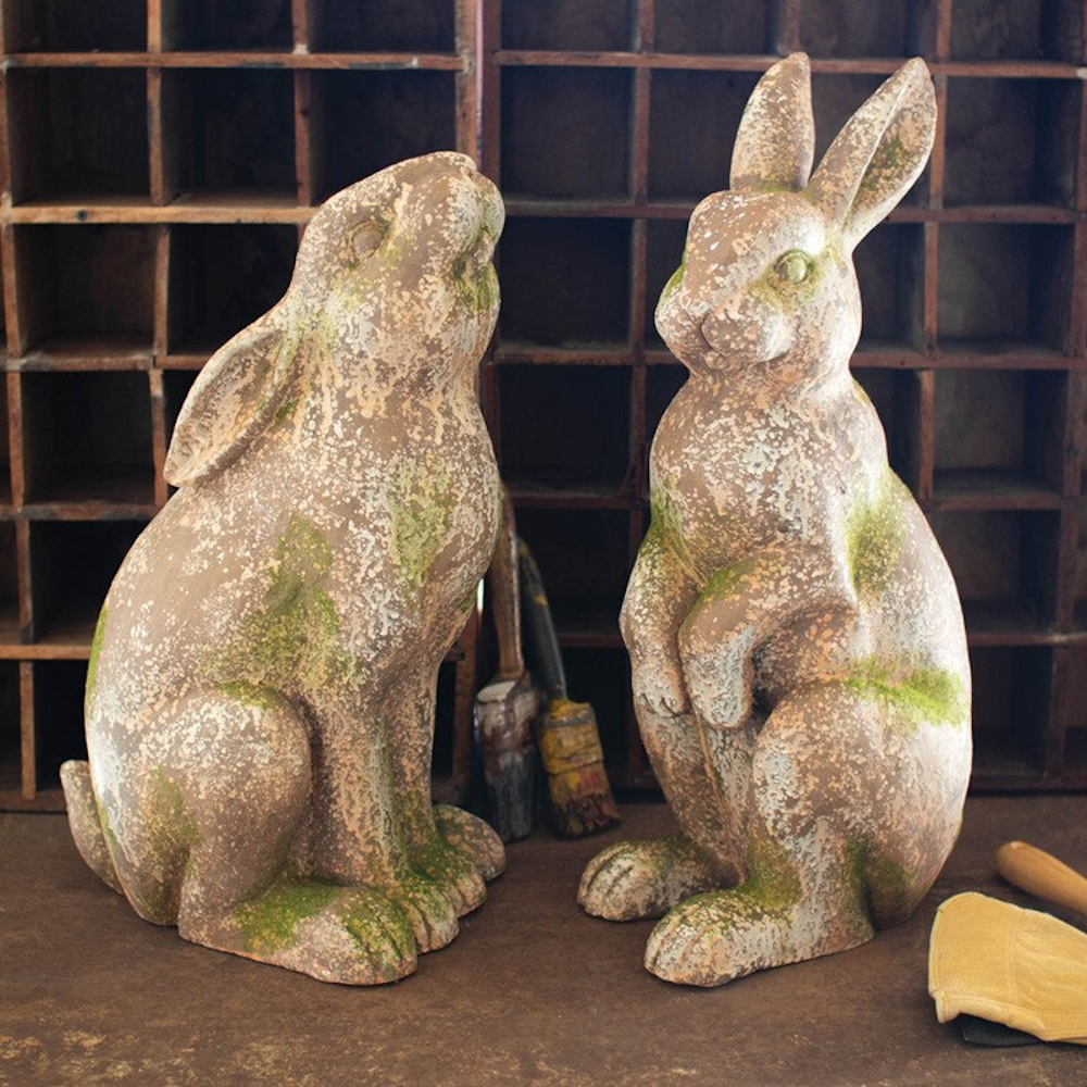 Two rustic faux-concrete rabbit statues with a weathered moss finish sitting in front of a vintage wooden cubby shelf.