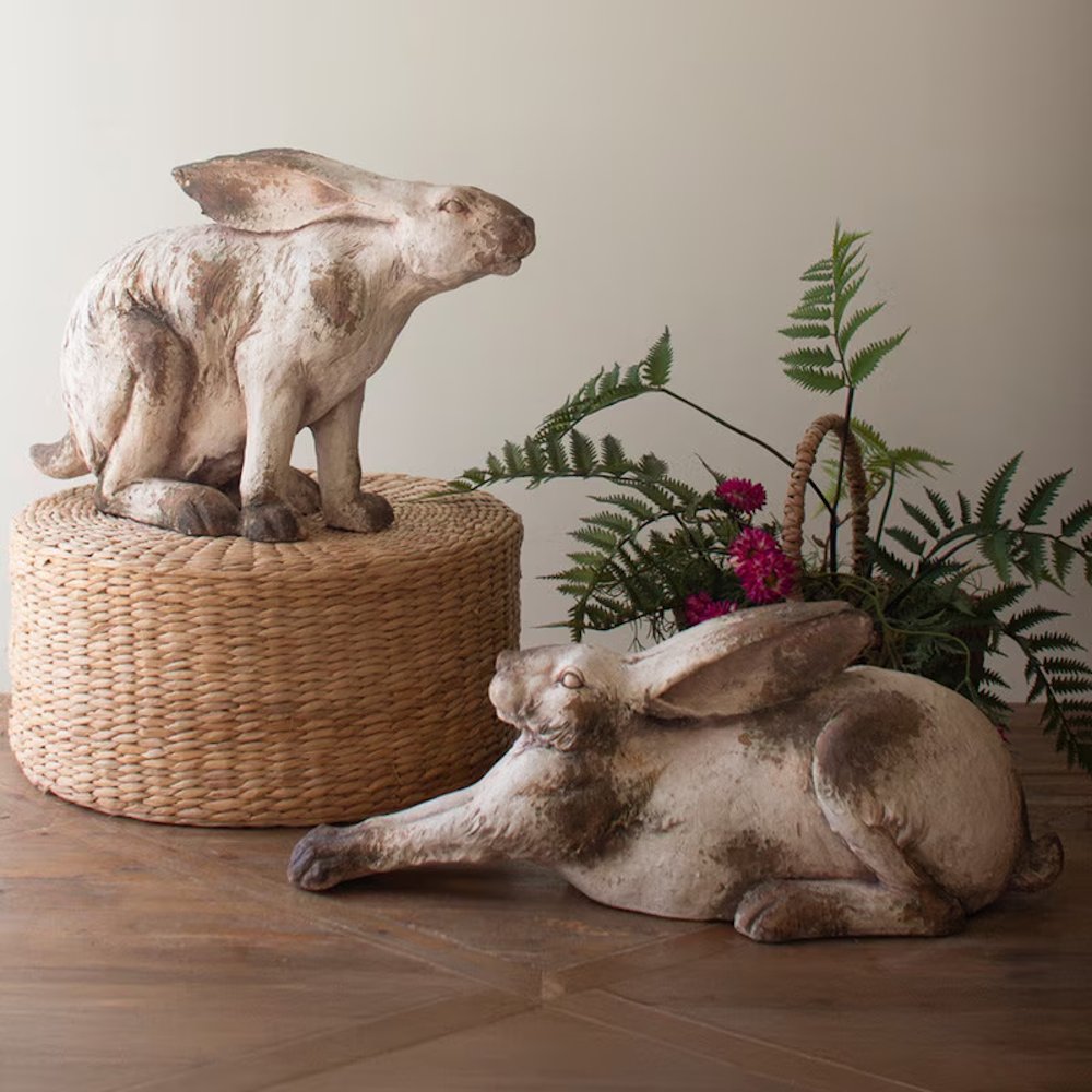 Two rustic faux concrete rabbit statues, one sitting and one reclining, displayed with greenery on a wooden table.
