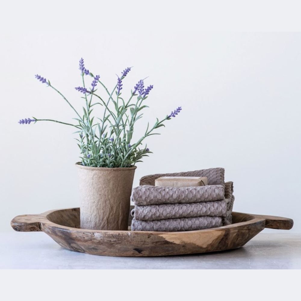 A potted lavender plant, a stack of grey waffle towels, and a bar of soap arranged on a rustic wooden tray.