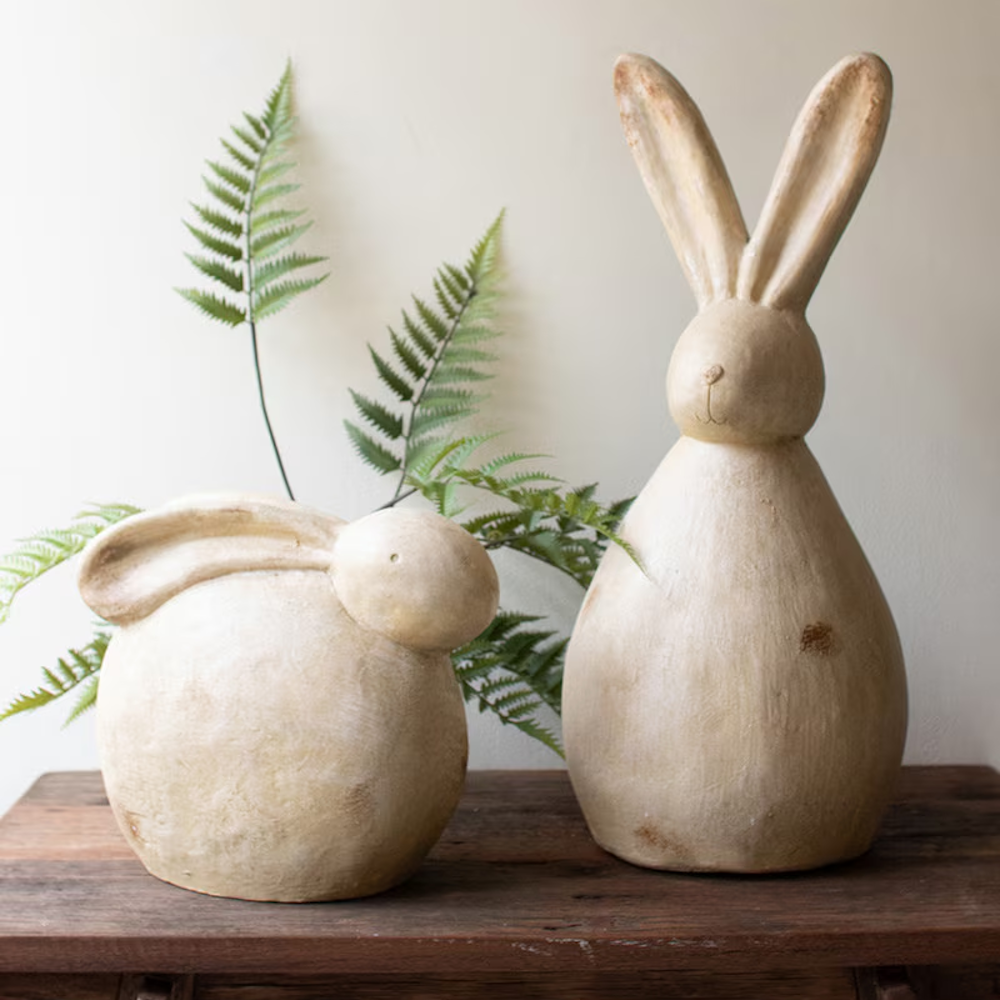 Two rustic faux stone rabbit sculptures of varying heights on a wooden surface with a green fern background.