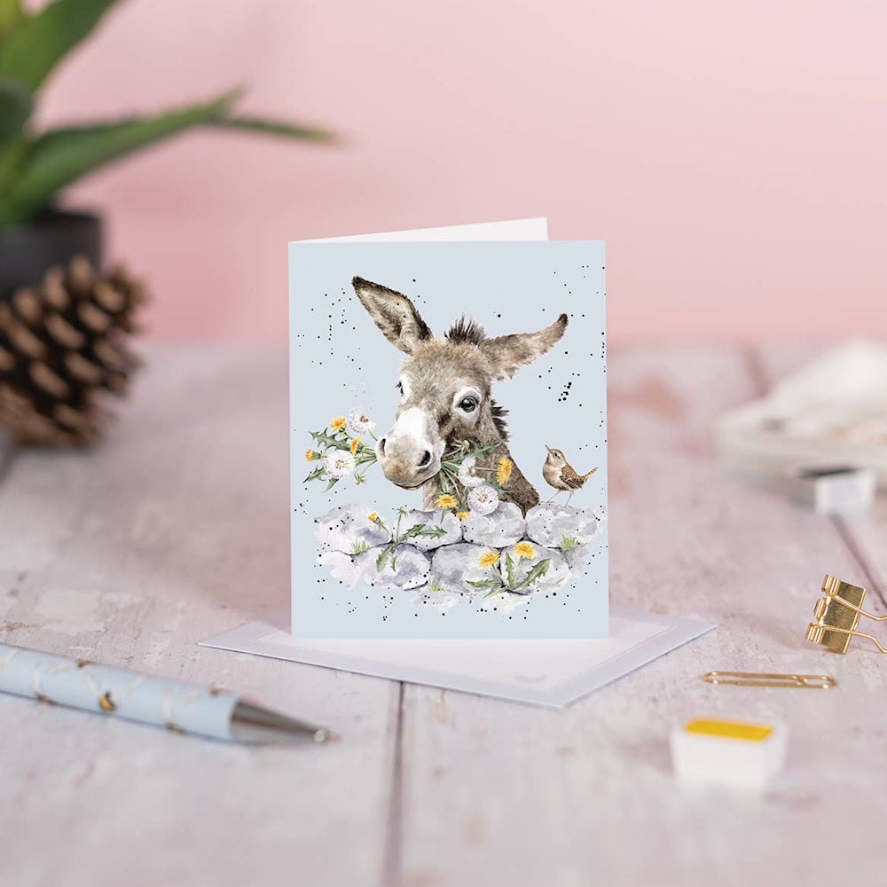 Watercolor donkey greeting card with dandelions and a bird, styled on a desk with a pen and gold clips.