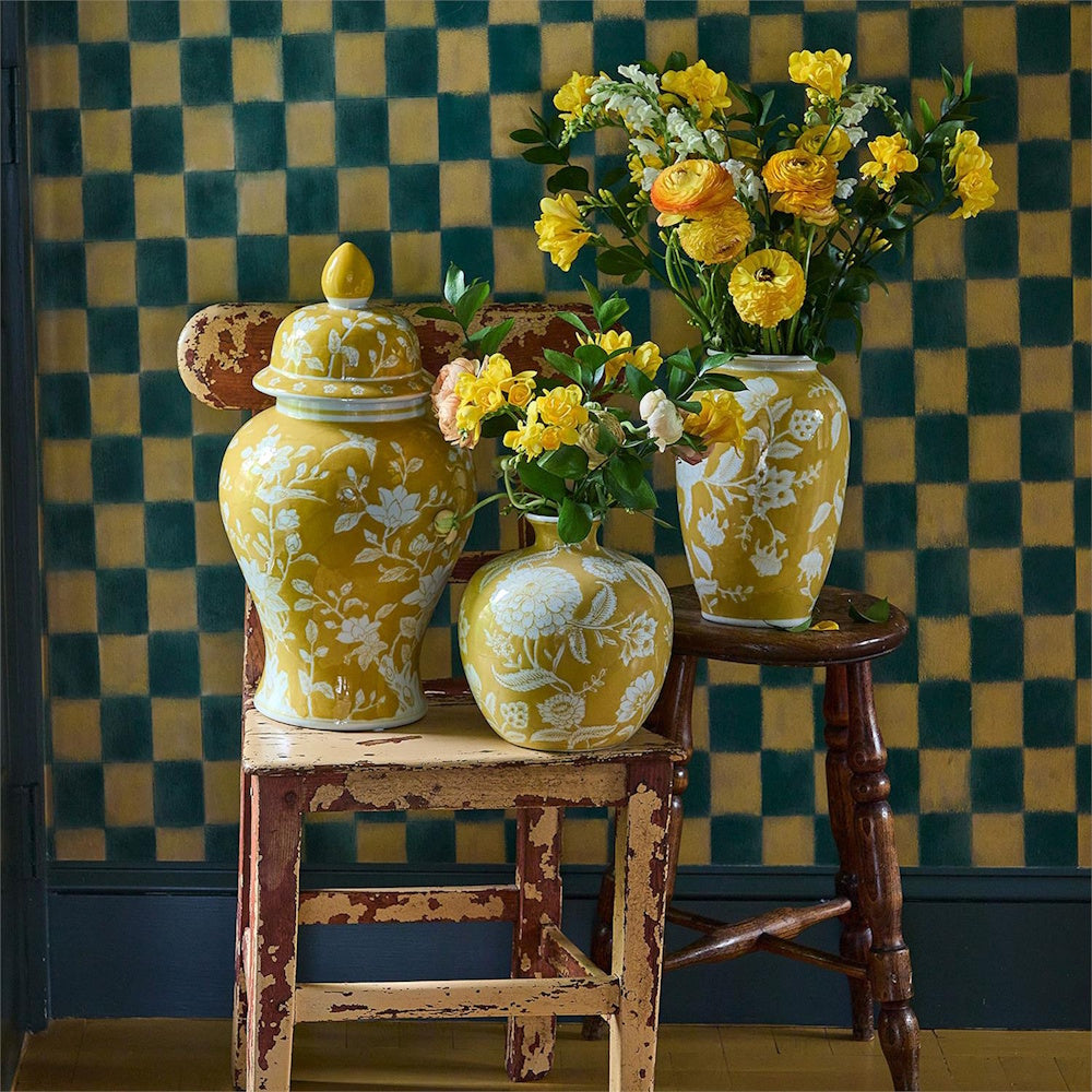Yellow floral ceramic vases with yellow flowers on rustic chairs against a green checkered wall.