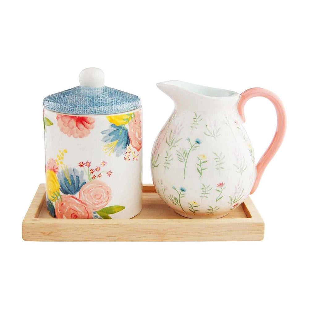 A floral ceramic cream and sugar set with a pink-handled pitcher and a lidded jar on a wooden tray.