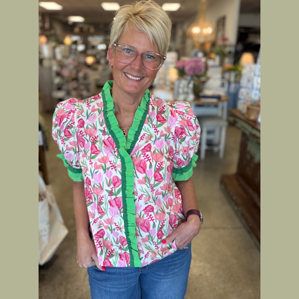 A woman with short blonde hair and glasses smiles wearing a floral puff-sleeve blouse with green ruffle trim.