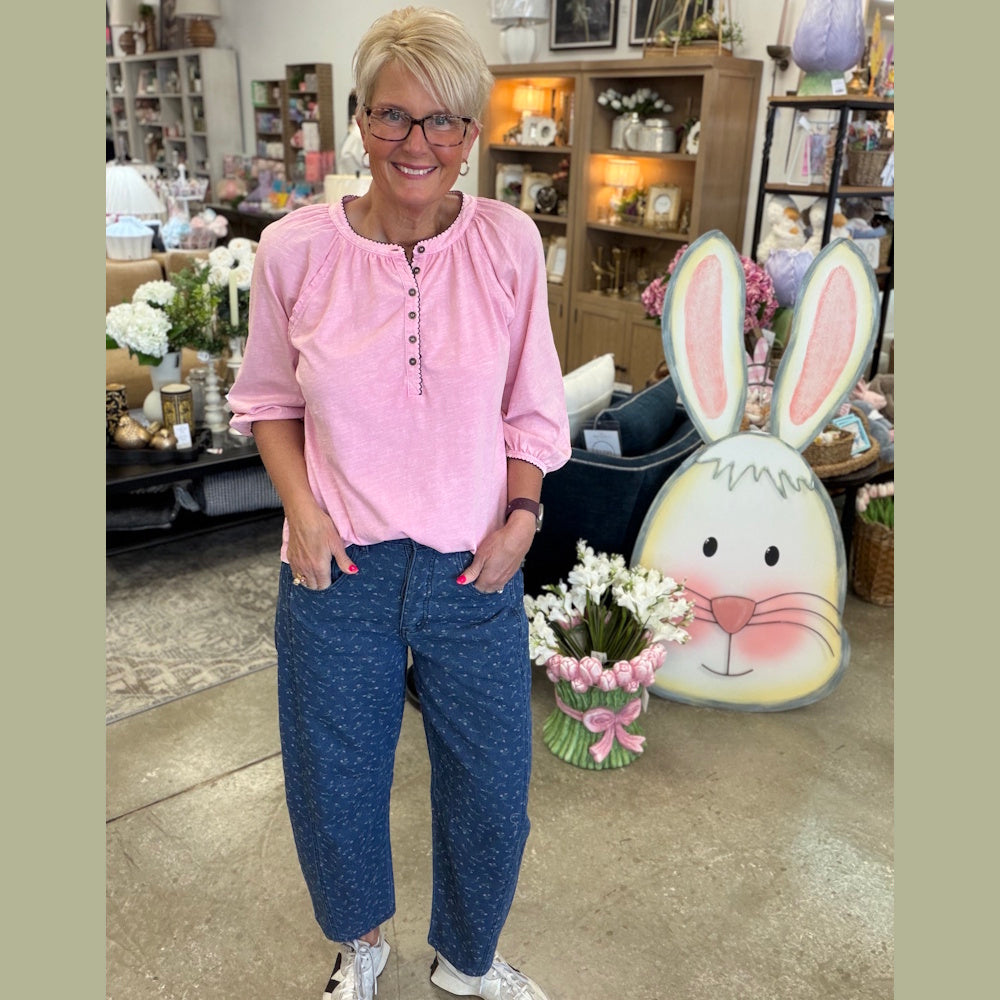 Woman wearing a pink henley top and floral-printed barrel denim pants in a store setting with Easter decor.