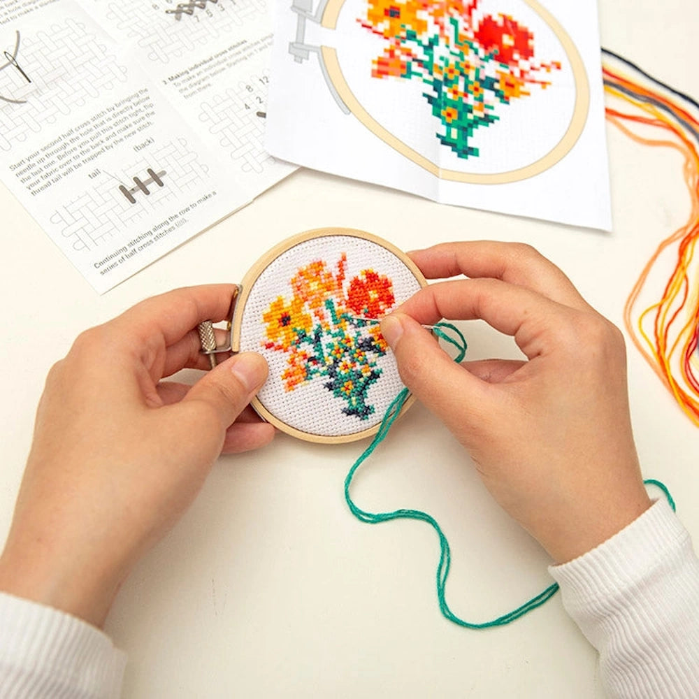 Hands stitching a mini floral bouquet cross-stitch in a wooden hoop with colorful embroidery thread and instructions.