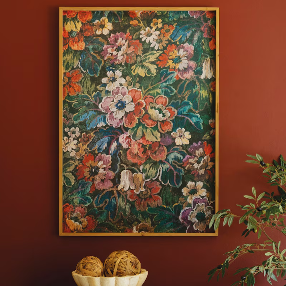 A framed floral tapestry art print on an earthy red wall, styled with a potted olive branch and decorative orb-filled bowl.