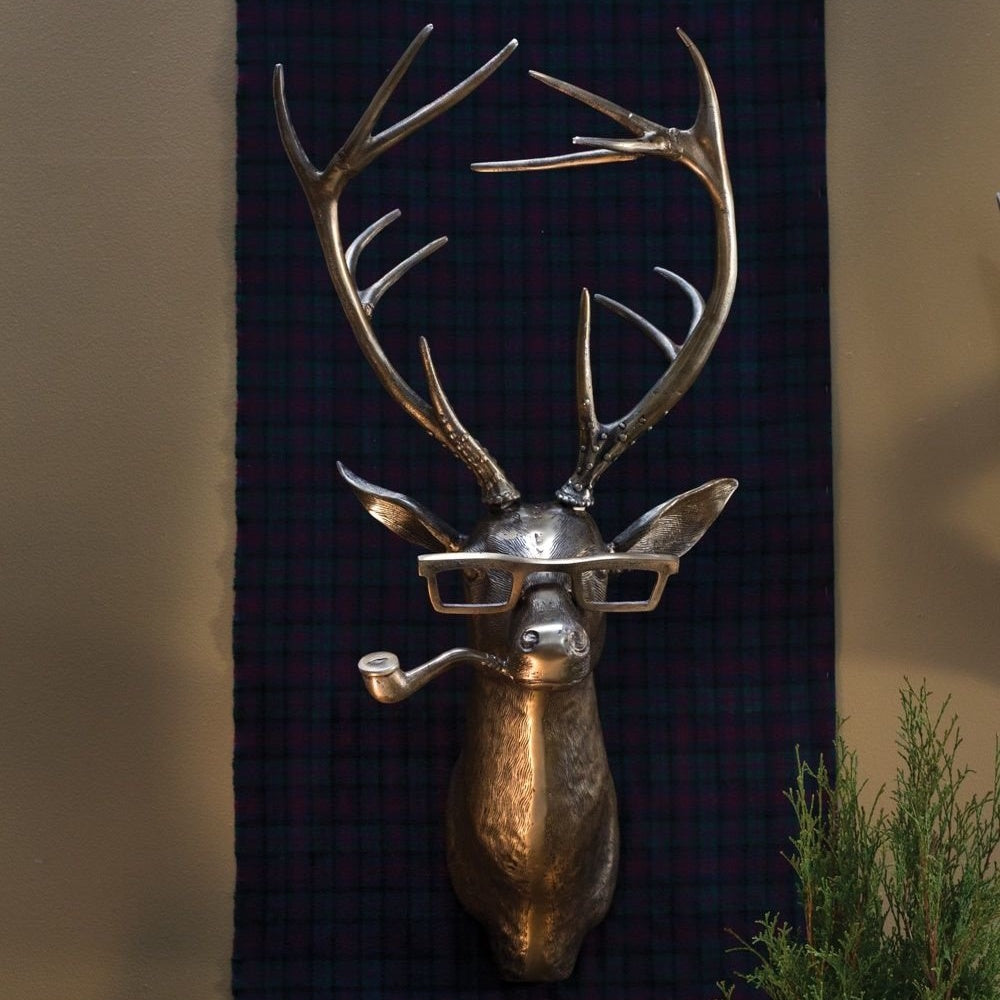 A metallic deer head wall mount wearing glasses and holding a pipe in its mouth, set against a plaid background.