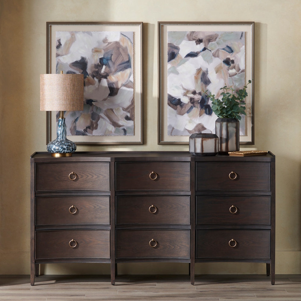 Dark wood 9-drawer dresser with gold ring pulls, table lamp, and abstract wall art in a modern neutral bedroom.
