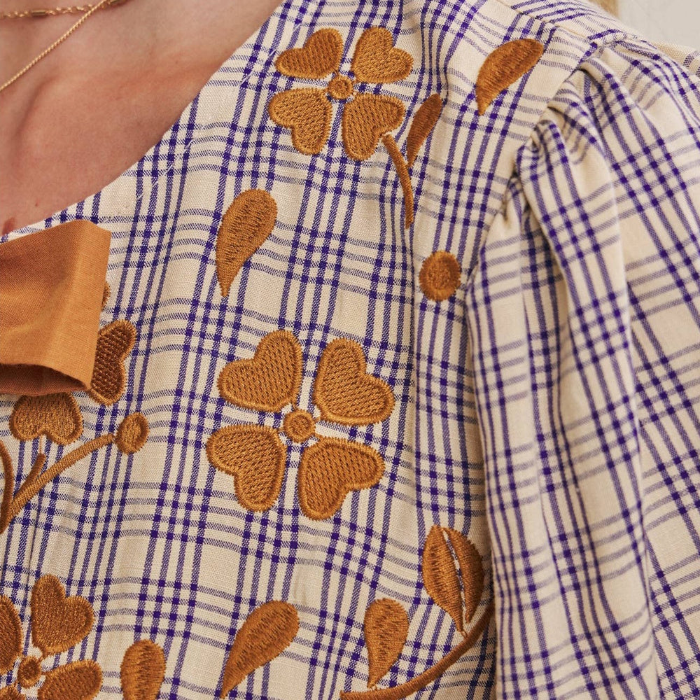 Close-up of a blue plaid blouse with gold floral embroidery, a brown bow detail, and a puff sleeve.