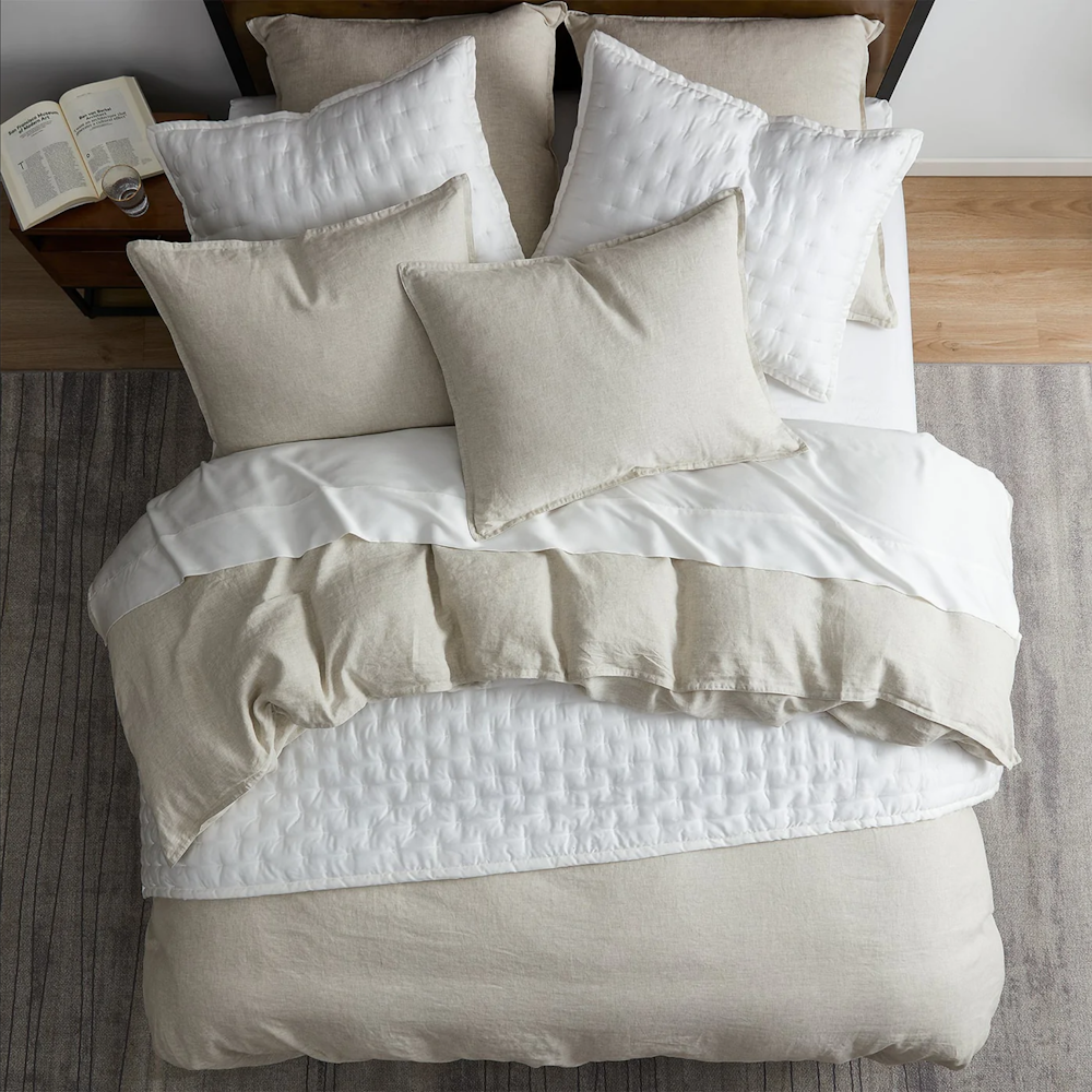 Top-down view of a bed with layered white and beige bedding, pillows, and a textured white quilt.