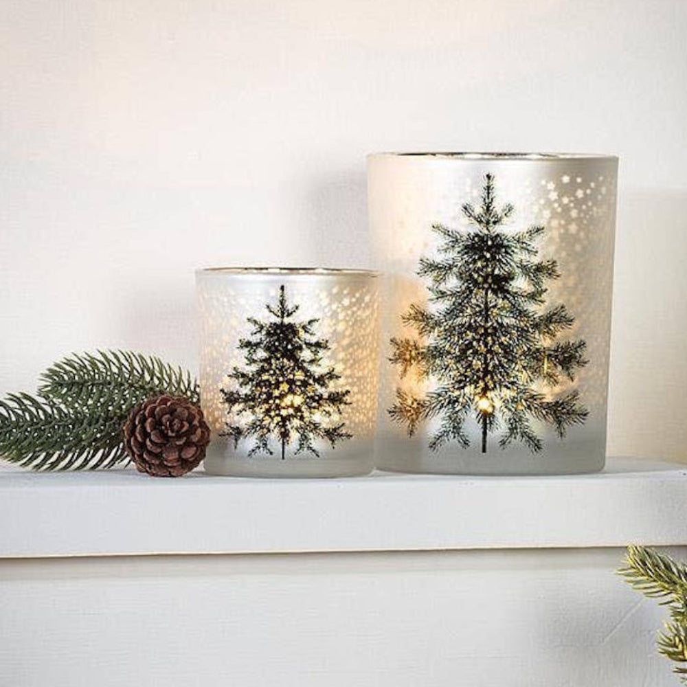 Two glowing frosted glass candle holders featuring pine trees and starry patterns on a mantle.