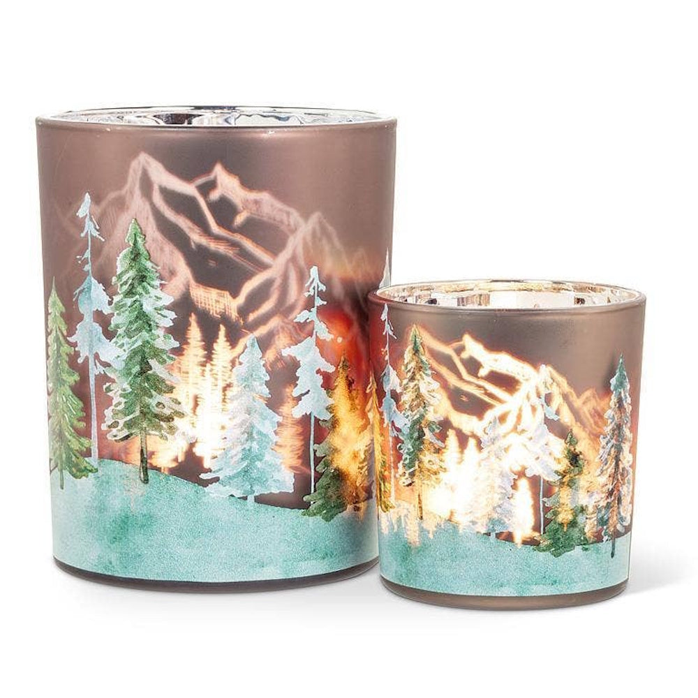 Two glass candle holders featuring frosty pine trees and glowing snowy mountain landscapes.