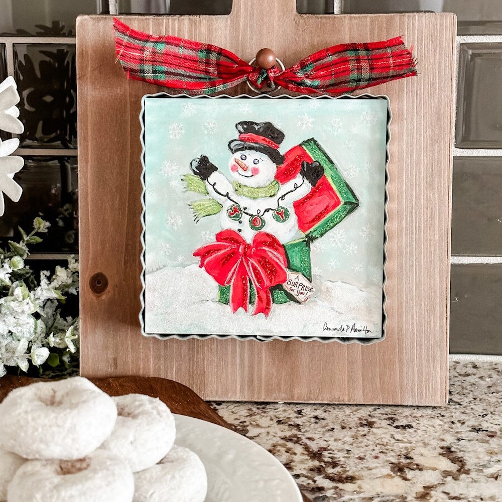 Front view of a square metal snowman art piece hanging on a wooden stand decorated with a red plaid ribbon.