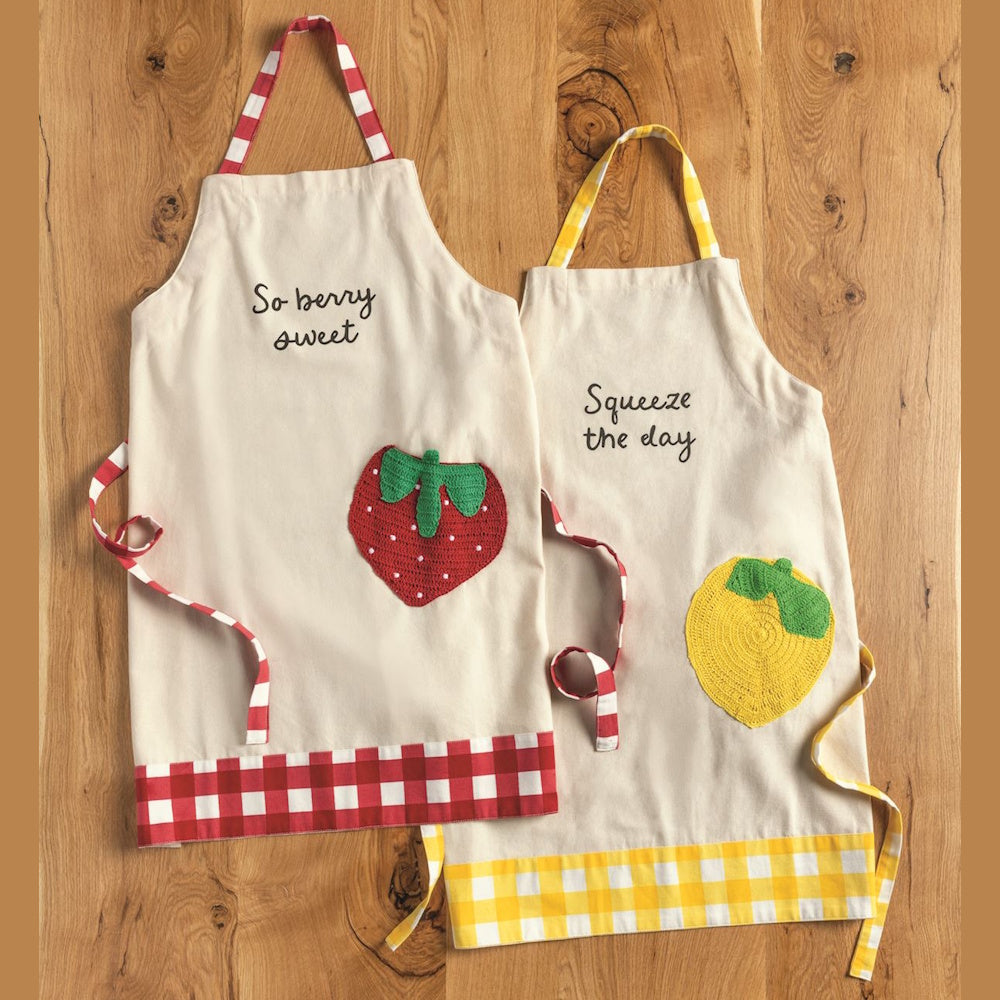 Two fruit-themed aprons with "So berry sweet" and "Squeeze the day" puns, strawberry and lemon pockets, and gingham trim.