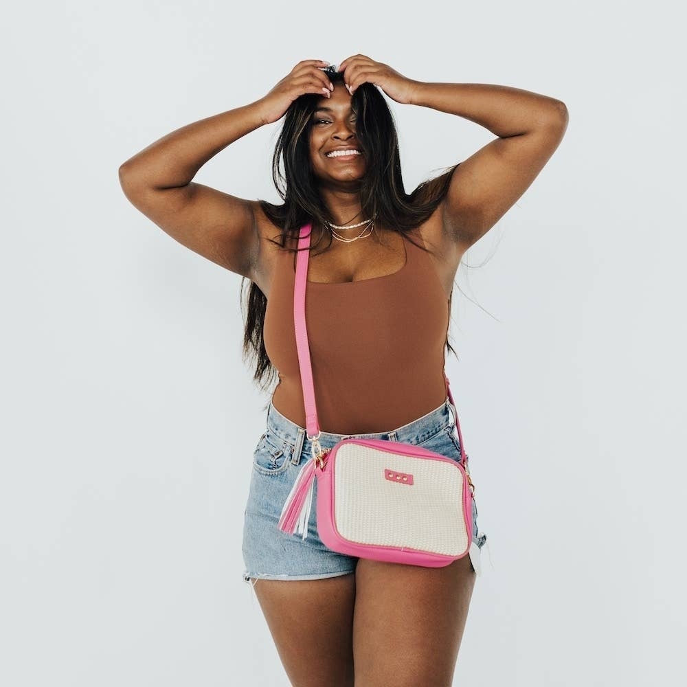 Smiling woman posing in denim shorts, a brown top, and a pink and woven straw crossbody bag.
