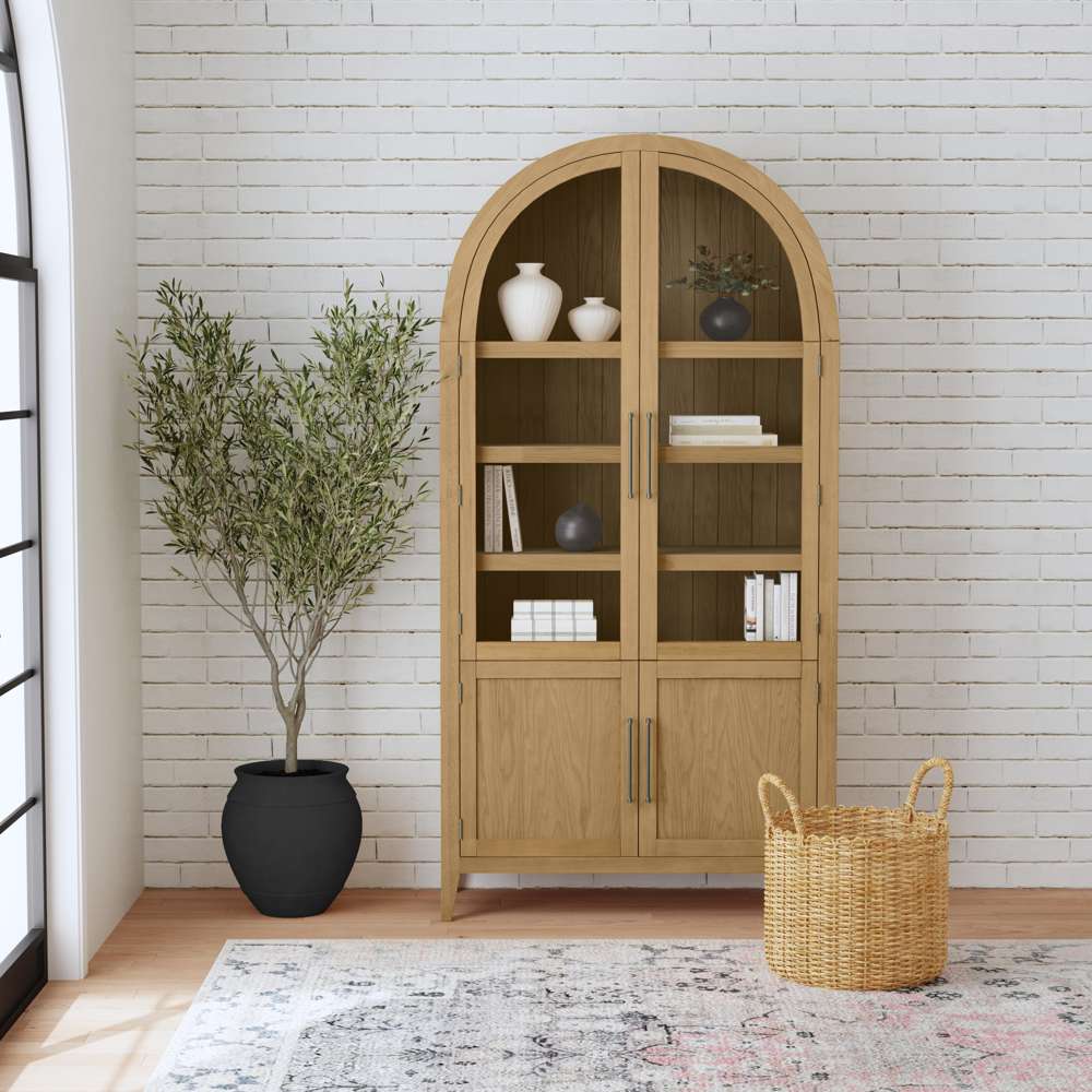 Arched light oak display cabinet with glass doors, styled with books and vases against a white brick wall.