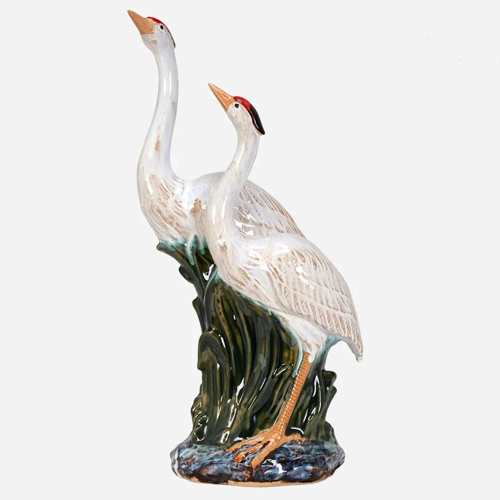 Ceramic figurine of two white cranes with red crowns, looking upward while standing in green marsh grass.