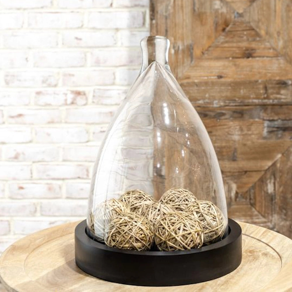 Bell-shaped glass cloche with a black wood base and decorative wicker spheres on a wooden table.