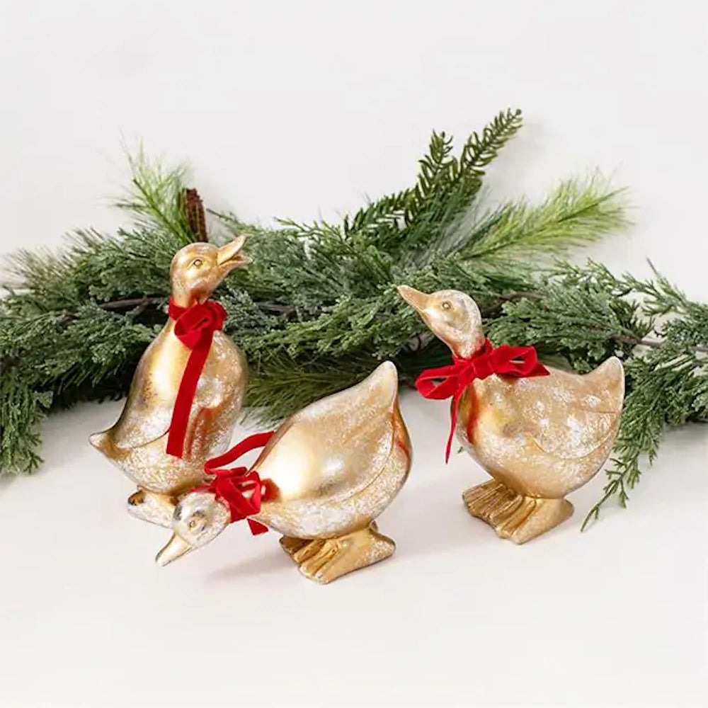 Three metallic gold duck figurines wearing red ribbon bows, set in front of evergreen pine branches.