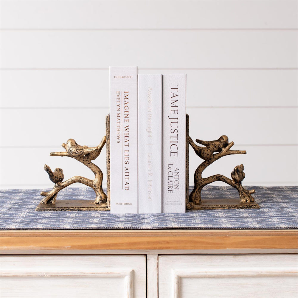 Gold-finish decorative bookends featuring birds on branches, holding white books on a blue patterned table runner.