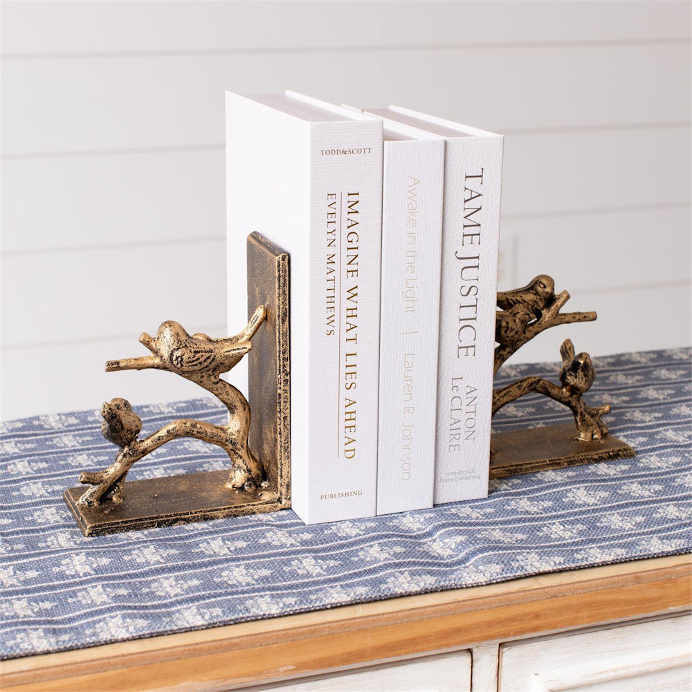 Gold finish bird on branch bookends holding three white books on a blue patterned table runner. Simple and direct.