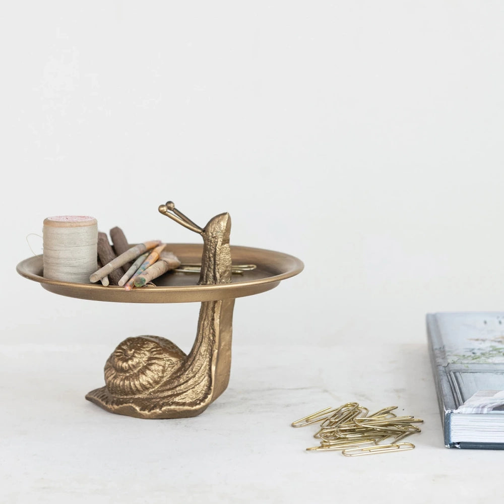 Gold-finish decorative metal tray on a snail-shaped stand, holding craft supplies and paperclips on a white surface.