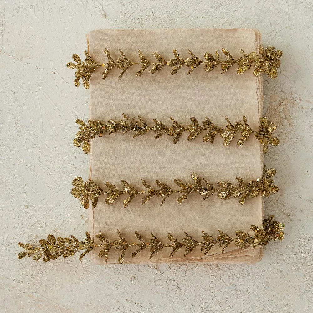 Gold finish faux boxwood garland with glitter, coiled on a textured neutral paper surface.