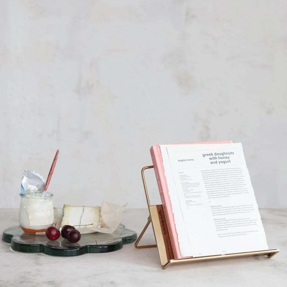 Modern gold bookstand with recipe book, yogurt jar, and cherries on a marble surface against a white textured wall.