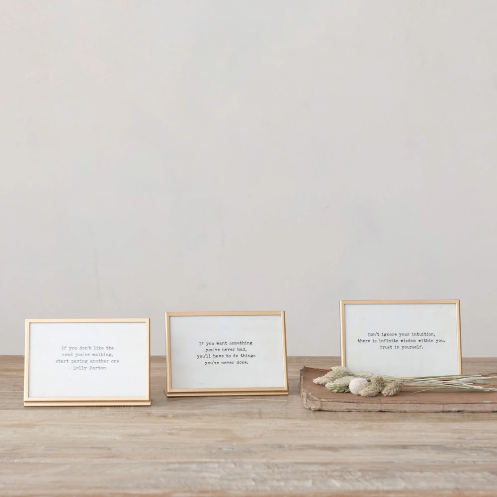 Three gold metal and glass picture frames with inspirational quotes on a rustic wooden table with dried flowers.