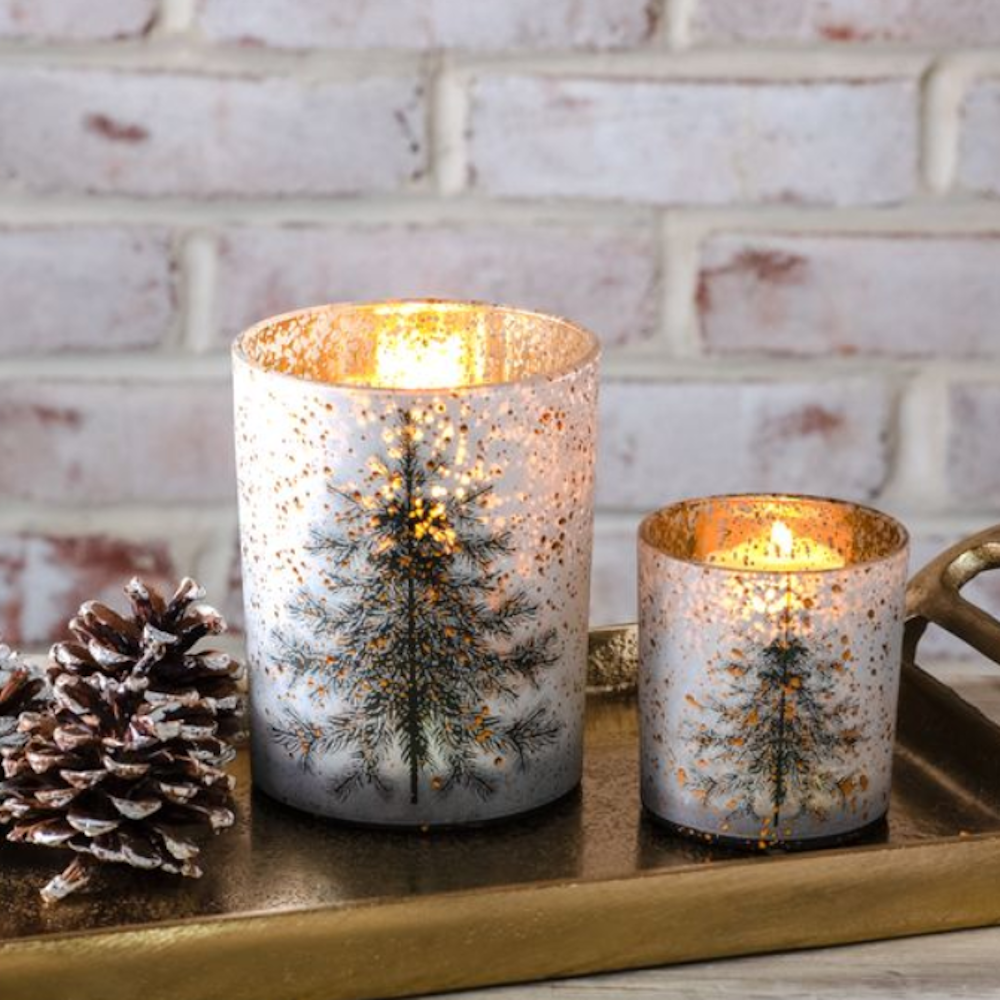 Pair of speckled glass hurricane candle holders with pine tree illustrations glowing on a tray.