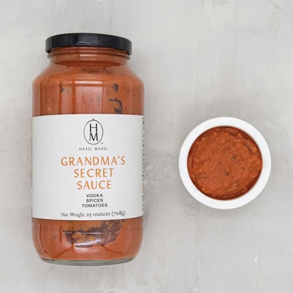 A jar and small bowl of Hazel Mazel Grandma’s Secret Sauce, a vodka tomato sauce, on a light grey background.