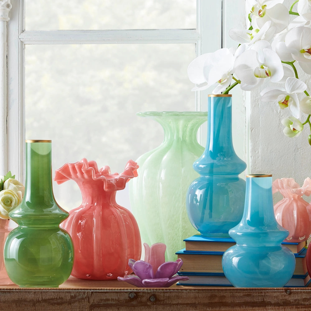 A collection of colorful glass bud vases in green, blue, and pink with gold rims on a shelf by a window.