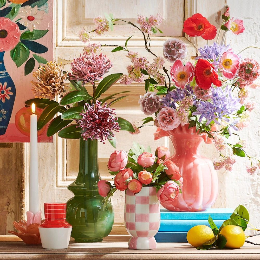 Colorful vases with flowers, a candle, and lemons on a table against a rustic wall and floral painting.