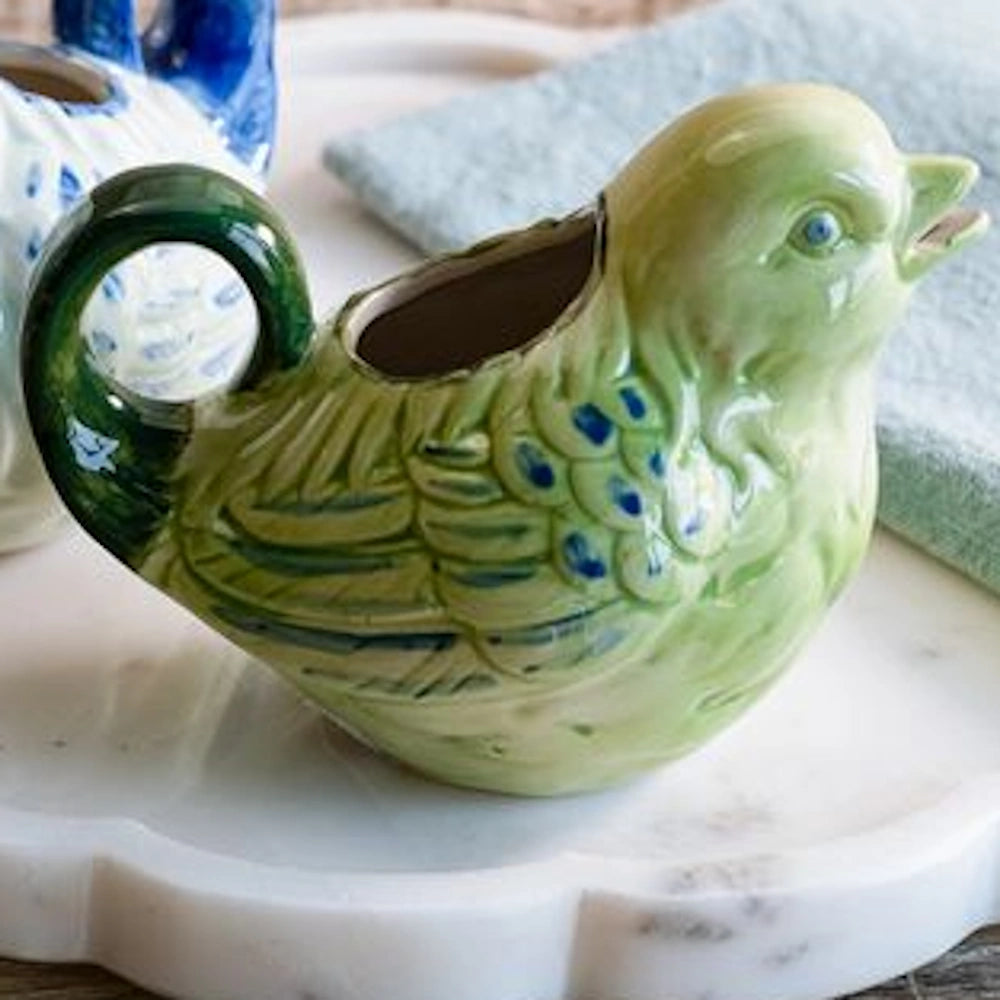 A decorative, light green ceramic bird creamer pitcher on a white marble surface next to a blue patterned napkin.