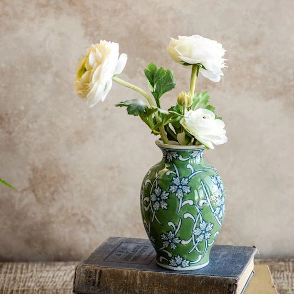 White ranunculus flowers in a green and blue floral ceramic vase resting on vintage books against a textured wall.