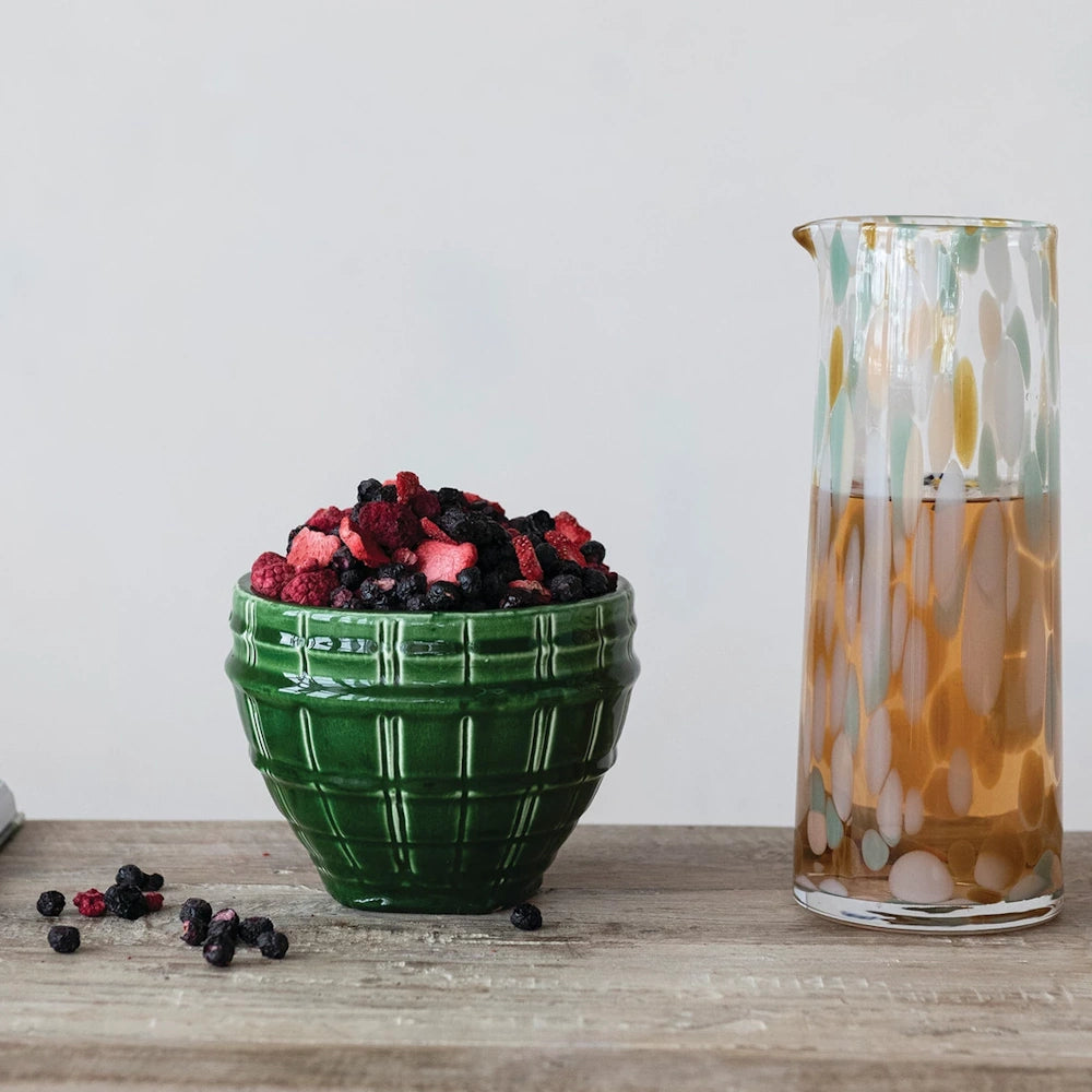 Green debossed stoneware planter filled with dried berries next to a spotted glass carafe on a wooden table.