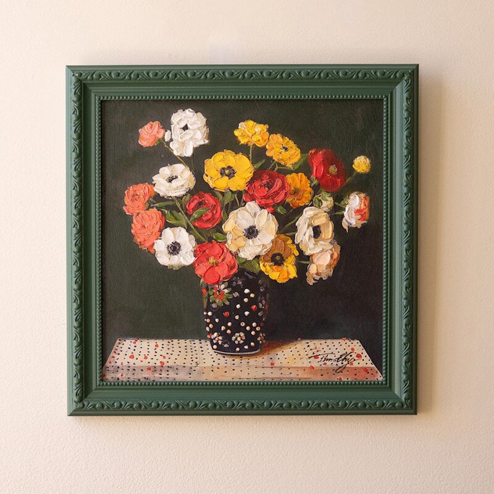 Still life oil painting of colorful flowers in a polka dot vase, featuring a decorative dark green frame on a beige wall.