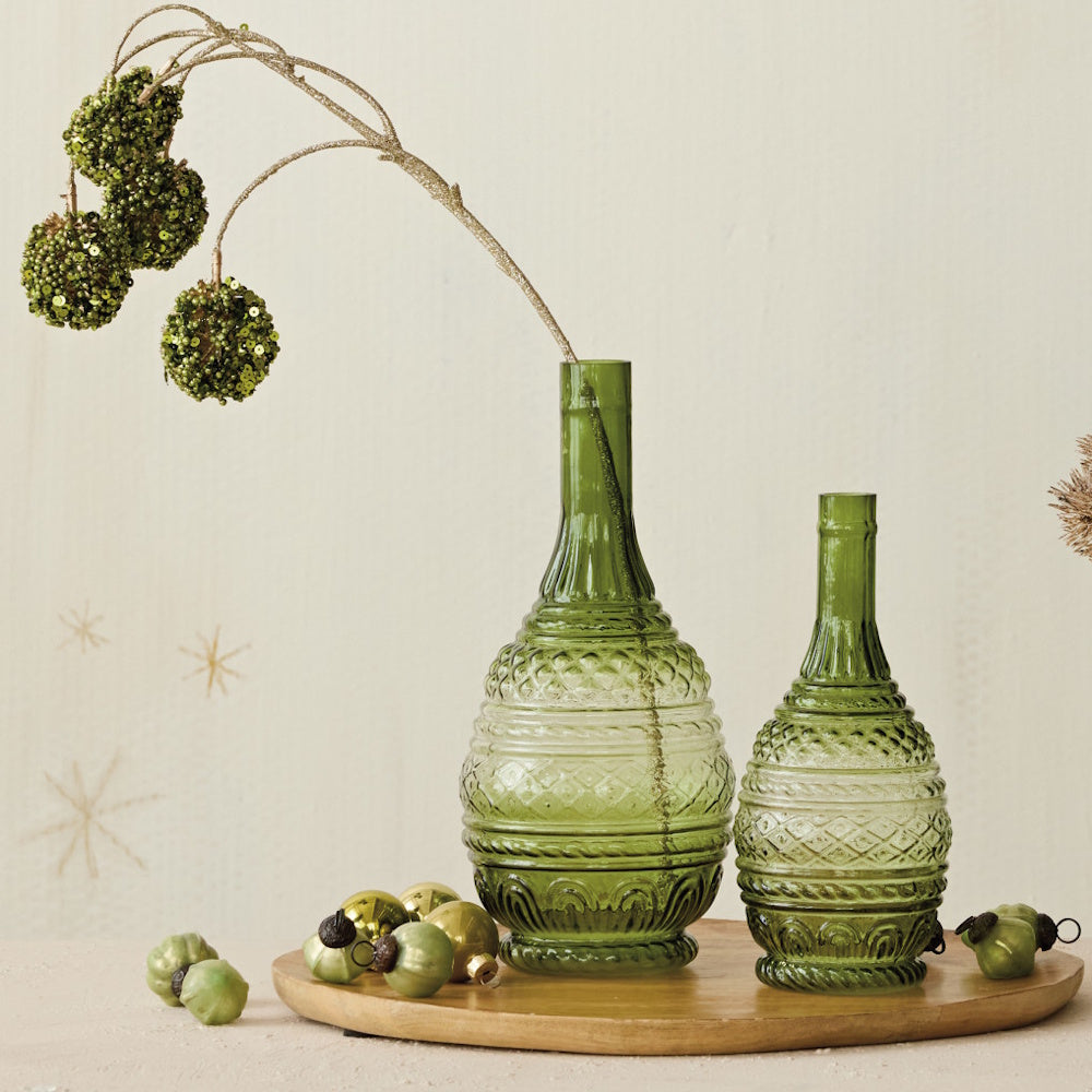 Two green embossed glass vases on a wooden tray with festive ornaments and glittery botanical stems.