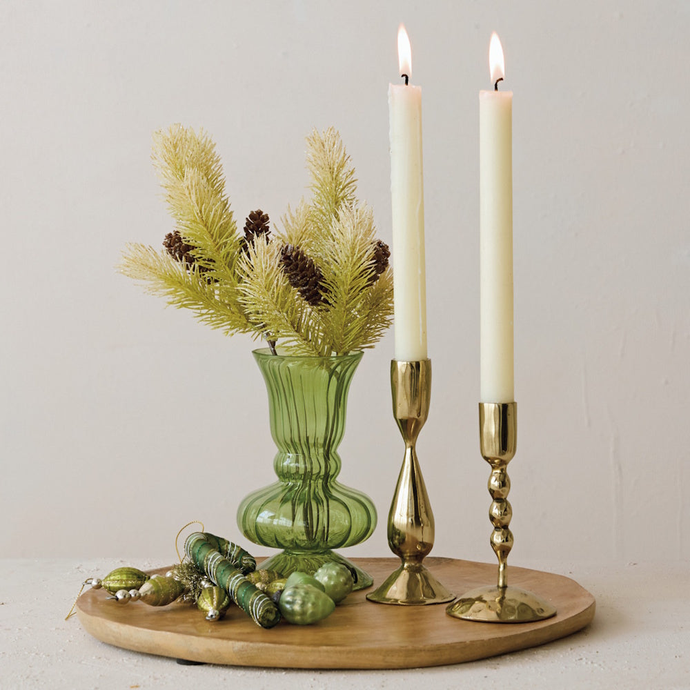 Green fluted glass vase with pine sprigs alongside brass candlesticks on a wooden tray.