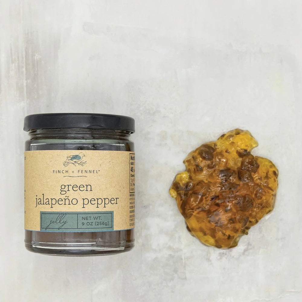 A jar of Finch + Fennel Green Jalapeño Pepper Jelly next to a scoop of golden jelly with pepper bits.