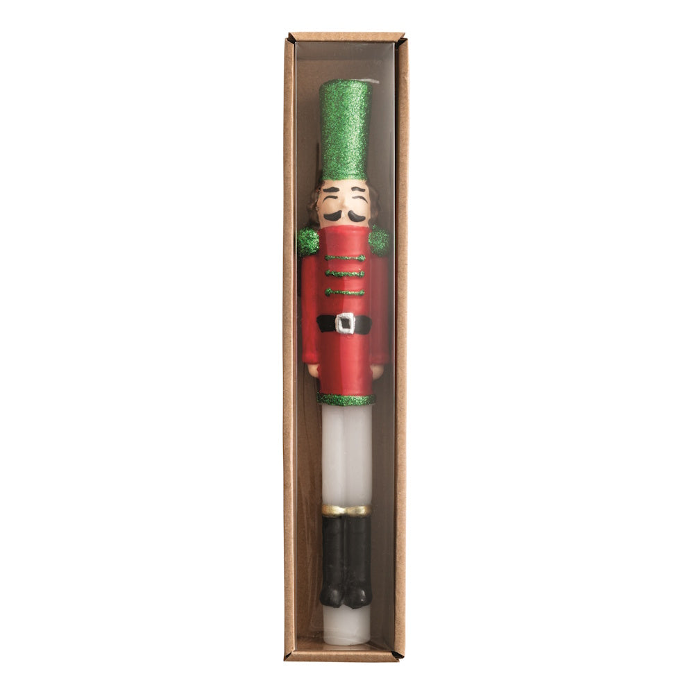 Front view of a red and white wax nutcracker taper candle with green glitter details in a boxed package.