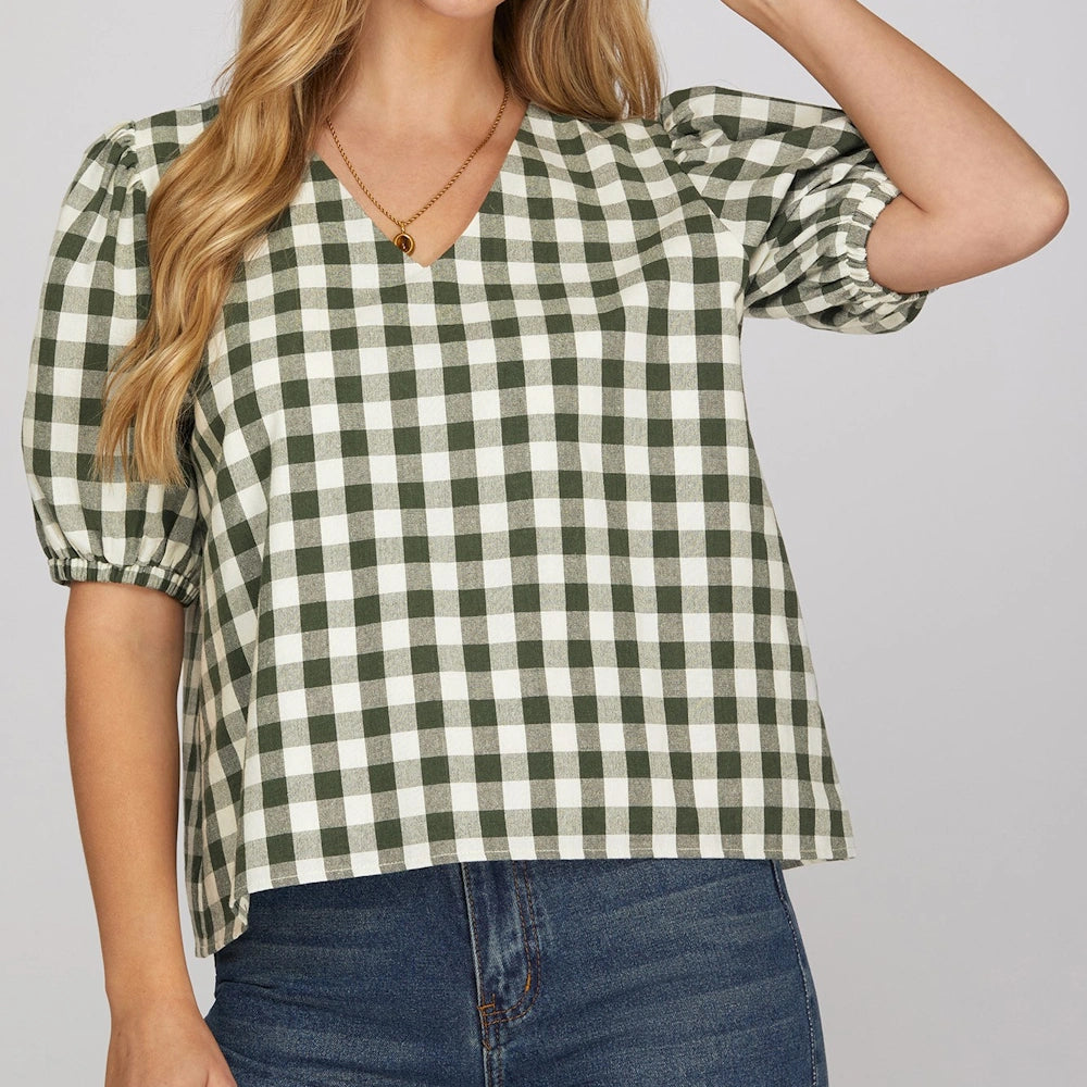 Woman wearing a green and white gingham checked v-neck top with puff short sleeves and blue jeans.