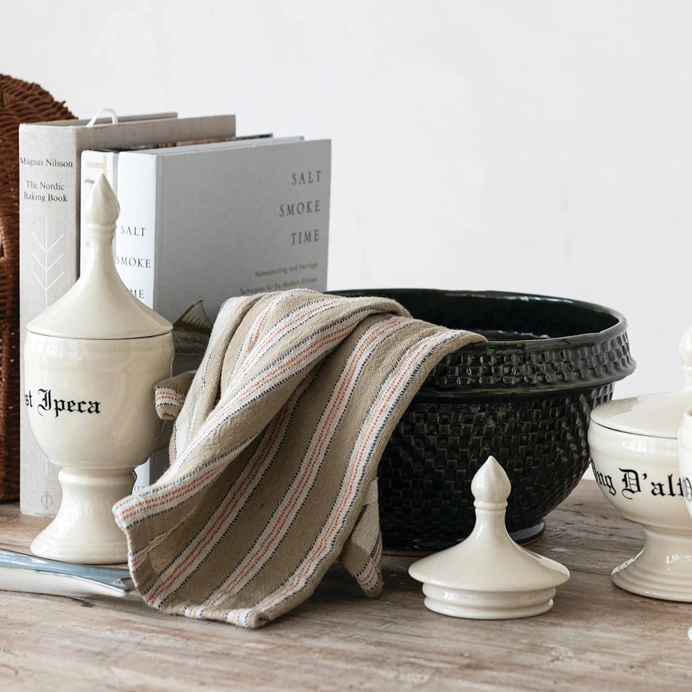Dark green basket weave stoneware bowl on a rustic table with striped cloth, cookbooks, and white apothecary jars.